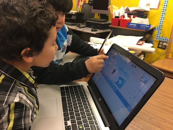 Two children are focused on working together at a laptop, with one pointing at the screen displaying a colorful, cartoon-like program. The setting suggests a classroom with various supplies and a bulletin board visible in the background.