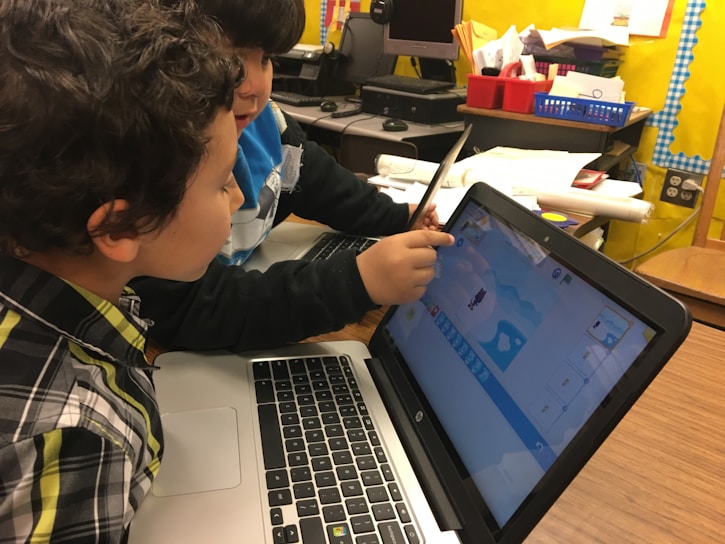 Two children are focused on working together at a laptop, with one pointing at the screen displaying a colorful, cartoon-like program. The setting suggests a classroom with various supplies and a bulletin board visible in the background.