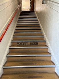 A staircase with wooden steps is leading upward, with a red railing on the left side. The words 'Poetry Room' are painted in black on one of the steps. The walls are covered with a textured pattern, and a light fixture is visible at the top of the stairs.