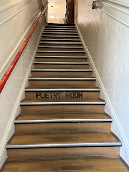 A staircase with wooden steps is leading upward, with a red railing on the left side. The words 'Poetry Room' are painted in black on one of the steps. The walls are covered with a textured pattern, and a light fixture is visible at the top of the stairs.