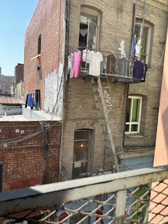 Clothes hang on a metal fire escape attached to an old brick building. The walls are weathered and display some graffiti. In the foreground, a rusted railing is visible. Multiple windows show various clothing items drying in the sun.