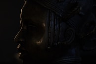 A dark silhouette of a detailed bronze sculpture depicting a face in profile view. The sculpture features intricate patterns and textures with visible lines and contours on the surface.