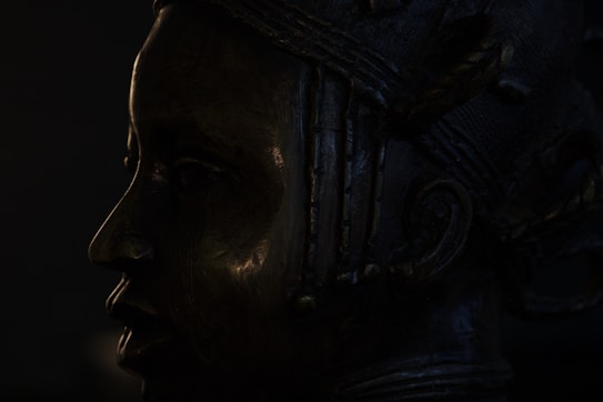 A dark silhouette of a detailed bronze sculpture depicting a face in profile view. The sculpture features intricate patterns and textures with visible lines and contours on the surface. Tim Burtom style
