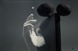 A monochrome composition featuring a hand reaching towards a small sphere. Two dark, spherical objects appear elevated and balanced on a vertical rod, casting shadows against a flat backdrop.