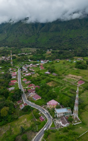 An aerial view of a rural area featuring a winding road lined with houses. The landscape is lush with greenery, and there are fields scattered around. Two tall communication towers are visible. In the background, large mountains partially obscured by low-hanging clouds provide a scenic backdrop.