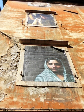 A weathered building facade displays two large, framed photographs. The top image features a person in a contemplative pose, sitting in soft light, while the bottom image shows a woman in a checkered veil gazing directly outward. The walls are aged, with visible cracks and peeling paint, giving the structure a rustic appearance.