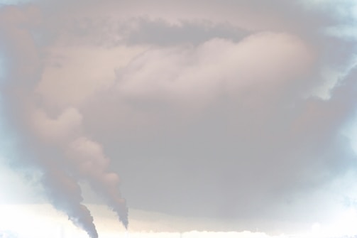Thick plumes of smoke rise from industrial chimneys into a cloudy sky, creating a hazy atmosphere.