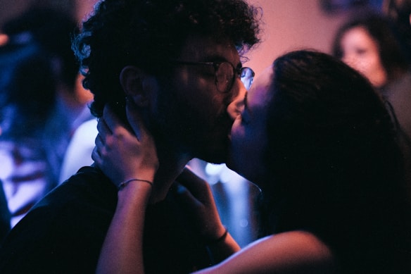 A couple is sharing an intimate kiss in a dimly lit setting, possibly a bar or club. The focus is on their faces with soft lighting creating a romantic atmosphere. The background is blurred, giving a sense of privacy and intimacy.