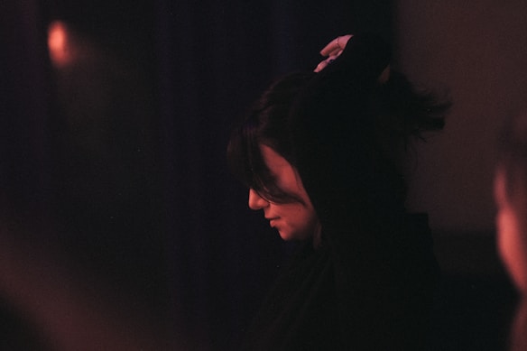 A person appears in a dimly lit environment, seen in profile with their head slightly tilted downward, as they adjust their hair. The lighting creates a warm glow, emphasizing shadows and contours.