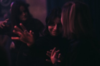 A dimly lit scene with people interacting closely. The focus is soft, contributing to a mysterious atmosphere. Subtle lighting highlights expressions and hand gestures, creating an intimate and somewhat enigmatic mood.