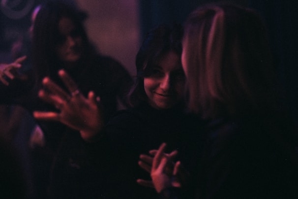 A dimly lit scene with people interacting closely. The focus is soft, contributing to a mysterious atmosphere. Subtle lighting highlights expressions and hand gestures, creating an intimate and somewhat enigmatic mood.