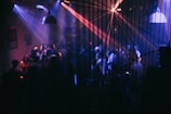 A dimly lit nightclub with purple, blue, and red lights projecting across the room. Numerous people are visible, dancing and socializing under the vibrant lights. The room includes hanging pendant lights and is adorned with various posters on the walls.