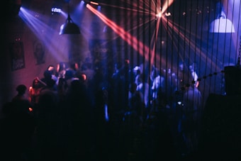 A dimly lit nightclub with purple, blue, and red lights projecting across the room. Numerous people are visible, dancing and socializing under the vibrant lights. The room includes hanging pendant lights and is adorned with various posters on the walls.