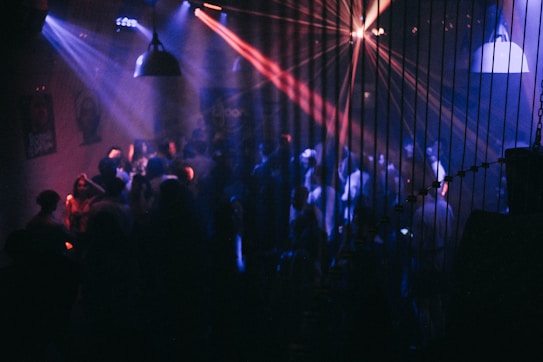 A dimly lit nightclub with purple, blue, and red lights projecting across the room. Numerous people are visible, dancing and socializing under the vibrant lights. The room includes hanging pendant lights and is adorned with various posters on the walls.