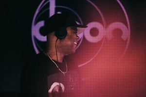 A person wearing headphones and a cap is smiling and appears to be enjoying themselves. The background features a circular design with indistinct text, and a reddish hue is noticeable in the foreground.