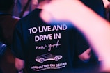 A person wearing a T-shirt featuring the text 'TO LIVE AND DRIVE IN new york' along with a graphic of a car and the words 'MANHATTAN CAR DEALER'. The scene includes blurred foreground elements and an ambient light setting, suggesting a nightlife or club environment.