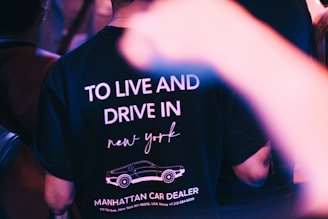 A person wearing a T-shirt featuring the text 'TO LIVE AND DRIVE IN new york' along with a graphic of a car and the words 'MANHATTAN CAR DEALER'. The scene includes blurred foreground elements and an ambient light setting, suggesting a nightlife or club environment.
