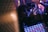 A person with curly hair and multiple rings is holding a smartphone, seemingly taking a photo or video. The background is dimly lit with people possibly dancing or socializing in a club-like environment, featuring vibrant, colored lighting. In the foreground, there is a digital grid or pad, likely a part of a DJ's equipment.