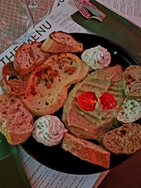 A black plate holds two types of dips, one garnished with cherry tomatoes and the other with chickpeas. Slices of crusty bread are arranged around the plate, along with swirls of a creamy topping. The setting includes a menu and glassware in the background.