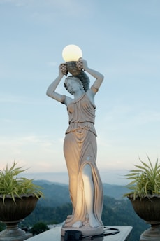 A classical statue of a woman holding a glowing orb above her head, set against a backdrop of mountains and a clear sky. The figure is elegantly draped in a flowing garment and flanked by potted plants.