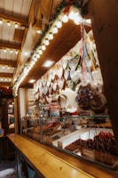 A market stall filled with various colorful sweets and treats. Gingerbread hearts with messages hang above a display of candies, candied apples, and chocolate-covered snacks. Soft yellow string lights and garlands with pine needles create a warm and festive atmosphere.