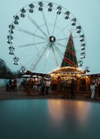 A large Ferris wheel towers over a festive scene with a Christmas tree adorned with lights and decorations. Below, people are gathered around wooden stalls, creating a bustling market atmosphere. The mood suggests a winter holiday celebration.