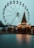 A large Ferris wheel towers over a festive scene with a Christmas tree adorned with lights and decorations. Below, people are gathered around wooden stalls, creating a bustling market atmosphere. The mood suggests a winter holiday celebration.
