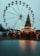 A large Ferris wheel towers over a festive scene with a Christmas tree adorned with lights and decorations. Below, people are gathered around wooden stalls, creating a bustling market atmosphere. The mood suggests a winter holiday celebration.
