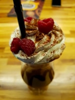 A glass of iced coffee topped with whipped cream, cocoa powder, and fresh raspberries. A black straw is inserted into the drink, and the glass stands on a wooden table. The rich texture and colors create an inviting and indulgent appearance.