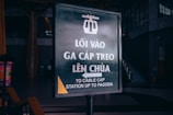 A signboard with Vietnamese and English text indicating the way to the cable car station leading to the pagoda. The sign features a cable car icon at the top and is situated indoors in a dimly lit area with some stairs visible in the background.