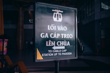 A signboard with Vietnamese and English text indicating the way to the cable car station leading to the pagoda. The sign features a cable car icon at the top and is situated indoors in a dimly lit area with some stairs visible in the background.
