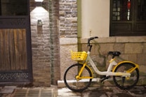 A yellow and white rental bicycle is parked against a textured stone wall, illuminated by a wall-mounted light above. The environment appears urban and the ground is slightly wet, suggesting recent rain or a damp atmosphere. The wall consists of a combination of stone blocks and brickwork, with a wooden door on one side.