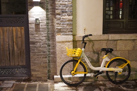 A yellow and white rental bicycle is parked against a textured stone wall, illuminated by a wall-mounted light above. The environment appears urban and the ground is slightly wet, suggesting recent rain or a damp atmosphere. The wall consists of a combination of stone blocks and brickwork, with a wooden door on one side.
