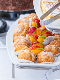 A variety of pastries arranged on a white platter. The assortment includes cream puffs dusted with powdered sugar and small tarts topped with colorful fruit slices like strawberries, kiwi, and oranges. Another plate holds slices of layered cake with a decorative drizzle on top. The setup exudes a fresh, inviting appeal.