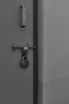 A sturdy metal door with a large rectangular handle and a sliding bolt that is secured by a round padlock. The surface of the door is plain and smooth, with a subtle texture.
