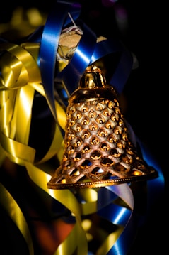 A decorative golden bell with intricate cut-out patterns is surrounded by metallic ribbons, primarily in blue and yellow, against a dark background.