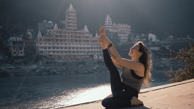 A person practicing yoga on a riverbank with a large temple-style building in the background. The person is engaged in a seated yoga pose, stretching one leg upwards with a serene expression. The scene is illuminated by soft morning or late afternoon light, casting gentle shadows.