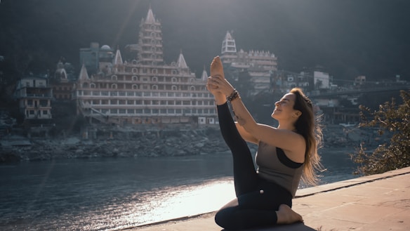 A person practicing yoga on a riverbank with a large temple-style building in the background. The person is engaged in a seated yoga pose, stretching one leg upwards with a serene expression. The scene is illuminated by soft morning or late afternoon light, casting gentle shadows.