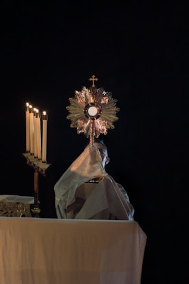 A religious ceremony with a figure holding a monstrance, an ornate object with a cross at the top, against a dark background. The figure is mostly hidden by a white garment. To the left, a candelabrum with several lit candles is visible, adding a warm glow to the scene.