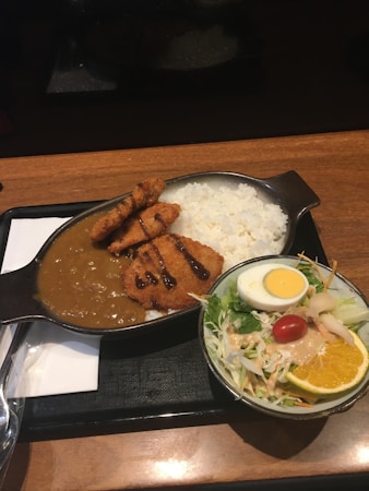 A meal is served on a black tray with a cast iron pan containing white rice, curry sauce, and breaded fried cutlets drizzled with dark sauce. Beside it, a small salad bowl includes sliced orange, cherry tomato, shredded cabbage, and a half boiled egg.