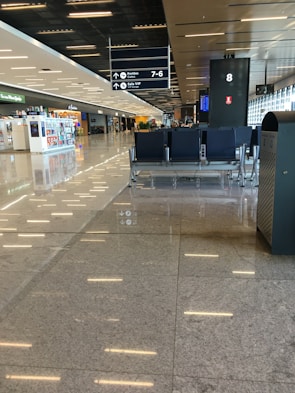 A spacious, modern airport terminal with a glossy, reflective floor. In the foreground, there is a row of empty black chairs. To the right, there is a garbage can. Further back, there are shops and restaurants along one side, and a row of large windows allowing natural light to enter. Overhead signs indicate directions to gates and a VIP lounge.