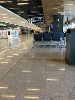 A spacious, modern airport terminal with a glossy, reflective floor. In the foreground, there is a row of empty black chairs. To the right, there is a garbage can. Further back, there are shops and restaurants along one side, and a row of large windows allowing natural light to enter. Overhead signs indicate directions to gates and a VIP lounge.