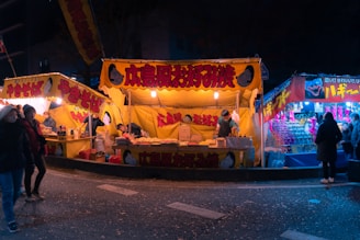 A vibrant night market scene with bright yellow stall canopies displaying Japanese food, likely yakisoba and okonomiyaki. Vendors are busy preparing food while people stroll by and shop at the stalls. The warm glow of lights contrasts with the cooler blue hues of nearby stalls.