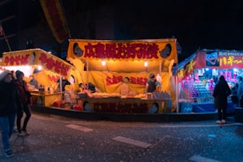 A vibrant night market scene with bright yellow stall canopies displaying Japanese food, likely yakisoba and okonomiyaki. Vendors are busy preparing food while people stroll by and shop at the stalls. The warm glow of lights contrasts with the cooler blue hues of nearby stalls.
