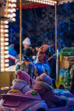 Children dressed in warm winter clothing sit together in a small, colorful ride shaped like a car. They are illuminated by the bright lights of what appears to be a carousel. In the background, other rides and blurred figures are visible, adding a festive atmosphere.