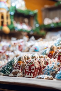 An assortment of intricately decorated gingerbread cookies is displayed, featuring designs such as gingerbread men, Christmas trees, snowflakes, and houses. These cookies are wrapped in transparent plastic, with icing in various colors like white, red, blue, and green adding festive detail. The background includes blurred greenery and warm lighting, suggesting a cozy, holiday setting.