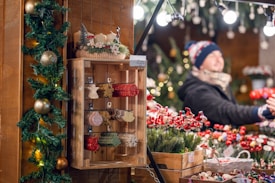 A festive market stall is decorated for the holiday season with greenery, golden ornaments, and a wooden display featuring various colorful ornaments. A person in the background is wearing winter clothing and appears to be smiling while interacting with visitors. The stall is filled with decorative items including small red and white mushrooms and other festive decorations. Bright lights add a warm and cheerful ambiance.