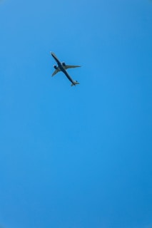 A commercial airplane flying through a clear blue sky, viewed from below. The aircraft appears to be mid-flight with its wings extended and engines visible.