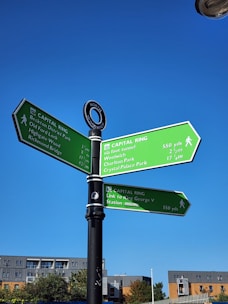 A signpost with three green directional panels indicating paths on the Capital Ring walkway. The panels provide distances to various locations such as Beckton District Park, Woolwich, and Crystal Palace Park. A symbol for North Woolwich is displayed above the signs. Below are buildings and a clear blue sky.