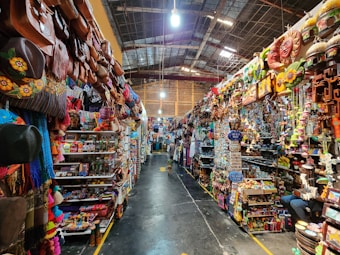 A vibrant, bustling indoor market stall filled with an array of colorful merchandise. The shelves are stocked with leather bags, hats, crafts, and various decorative items. The scene is lively, with bright colors and diverse items creating a visually rich environment.